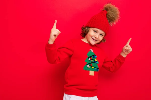 Pojke modell på en röd bakgrund, porträtt av en cool blondin med lockar i en tröja med en julgran — Stockfoto