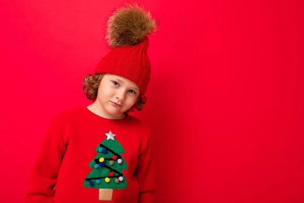 Cool blond unge i varm hatt och tröja med julgran på röd bakgrund lurar runt, jul koncept — Stockfoto