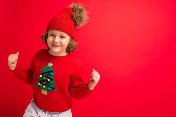 Niño rubio fresco en sombrero caliente y suéter con árbol de Navidad sobre fondo rojo tonteando alrededor, concepto de Navidad — Foto de Stock