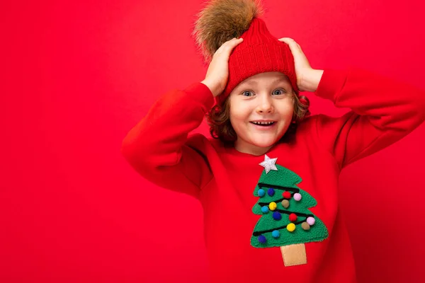 Adolescente fresco en un suéter rojo de Navidad jugando alrededor contra el fondo de una pared roja, un sombrero caliente y un suéter con un árbol de Navidad — Foto de Stock