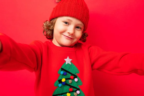 Lindo chico rubio en sombrero caliente y suéter de Navidad sobre fondo rojo con sonrisa en su cara — Foto de Stock