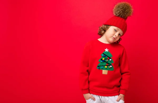 Niño rubio fresco en sombrero caliente y suéter con árbol de Navidad sobre fondo rojo tonteando alrededor, concepto de Navidad — Foto de Stock