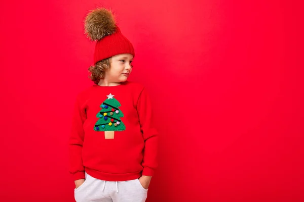 Δροσερό έφηβος σε ένα κόκκινο χριστουγεννιάτικο πουλόβερ χαζολογώντας γύρω από το φόντο ενός κόκκινου τοίχου, ένα ζεστό καπέλο και ένα πουλόβερ με ένα χριστουγεννιάτικο δέντρο — Φωτογραφία Αρχείου