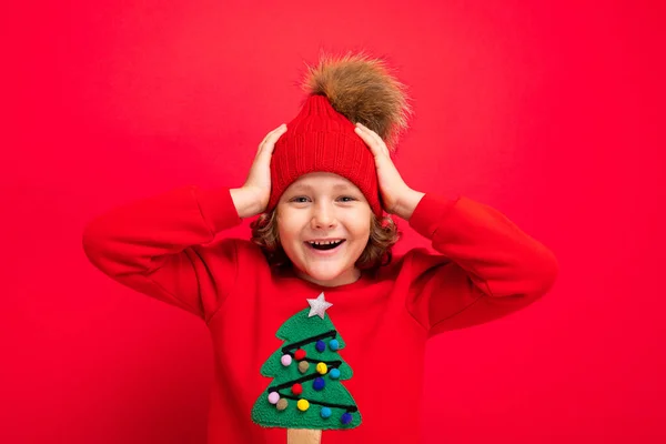 Adolescente fresco en un suéter rojo de Navidad jugando alrededor contra el fondo de una pared roja, un sombrero caliente y un suéter con un árbol de Navidad — Foto de Stock