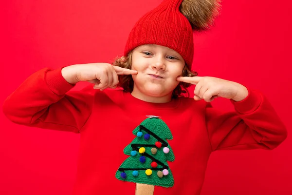 Modelo de niño sobre un fondo rojo, retrato de una rubia guay con rizos en un suéter con un árbol de Navidad — Foto de Stock