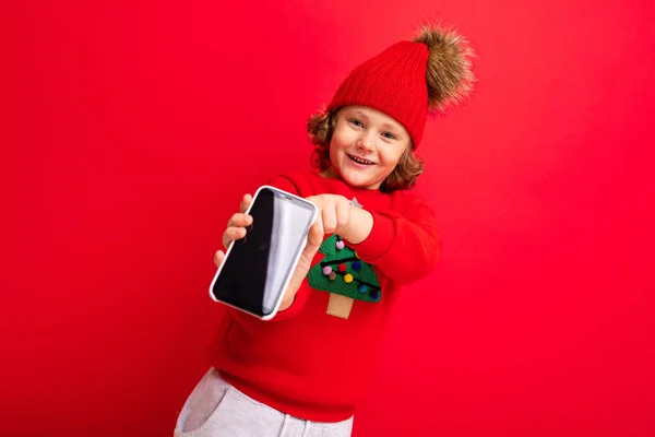 En ung man med en smartphone i händerna mot bakgrund av en röd vägg, i en stickad hatt och en tröja med en julgran. — Stockfoto