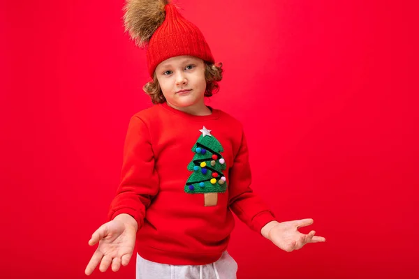 Mignon garçon blond en chapeau chaud et pull de Noël sur fond rouge avec sourire sur son visage — Photo