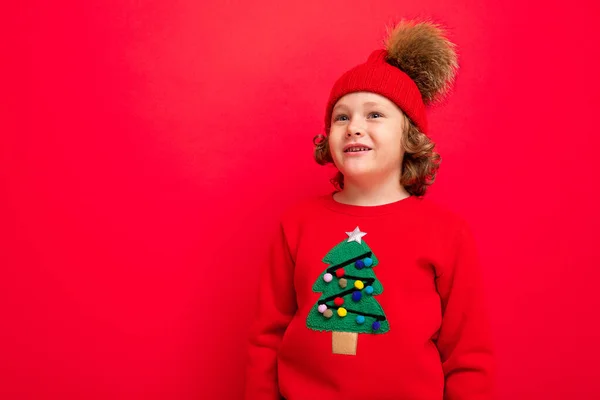 Joyeux garçon drôle sur un fond rouge dans un chapeau chaud et pull avec un arbre de Noël — Photo