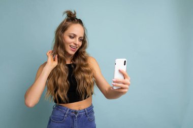 Çekici, genç, gülümseyen, mutlu bir kadın cep telefonu tutuyor ve kullanıyor. Selfie çekiyor. Duvar arkasından izole edilmiş şık kıyafetler giyiyor.