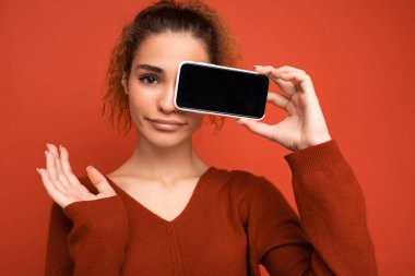 Kırmızı arka planda koyu kırmızı süveter giyen genç bir kadının yakın plan fotoğrafı. Elinde akıllı telefon, elinde de fotokopi alanı olan bir cep telefonu ekranı var.