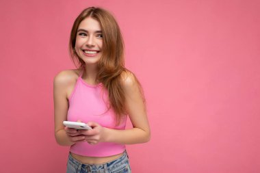 Gülümseyen, güzel görünümlü, pembe bluz giyen genç bir kadının fotoğrafı. Pembe arka planda izole edilmiş, boş bir alanda el ele tutuşmuş ve cep telefonuyla mesajlaşan bir kadın.