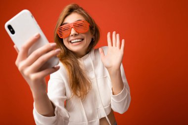 Çekici, genç, gülümseyen, mutlu bir kadın cep telefonu tutuyor ve kullanıyor. Selfie çekiyor. Duvar arkasından izole edilmiş şık kıyafetler giyiyor.