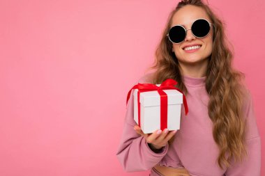 Çekici, pozitif gülümseyen genç, sarışın bir kadın. Renkli arka plan duvarından izole edilmiş, günlük moda kıyafetler giymiş, elinde hediye kutusuyla kameraya bakıyor.