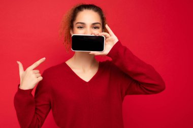 Güzel, genç bir kadının resmi. Günlük kıyafetlerini giymiş. Arka planda izole bir şekilde duruyor. Akıllı telefonu elinde tutuyor ve elinde boş bir ekran görüntüsü var.