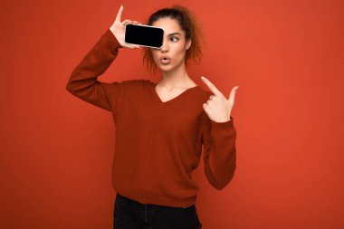 Kırmızı arka plan üzerinde koyu kırmızı süveter giyen, elinde akıllı telefon tutan ve fotokopi için cep telefonu ekranı olan mutlu, genç bir kadının fotoğrafı.
