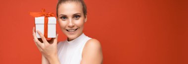 Güzel, pozitif gülümseyen sarışın bir kadının panoramik bir fotoğrafı. Kırmızı arka plan duvarında izole edilmiş, beyaz bluzlu, elinde hediye kutusu olan ve kameraya bakan bir kadın.