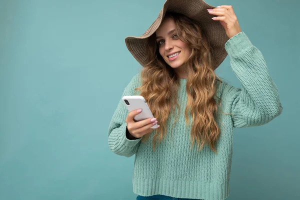 Beautiful happy smiling young blonde woman wearing blue sweater and hat standing isolated over blue background surfing on the internet via phone looking at camera — Fotografia de Stock