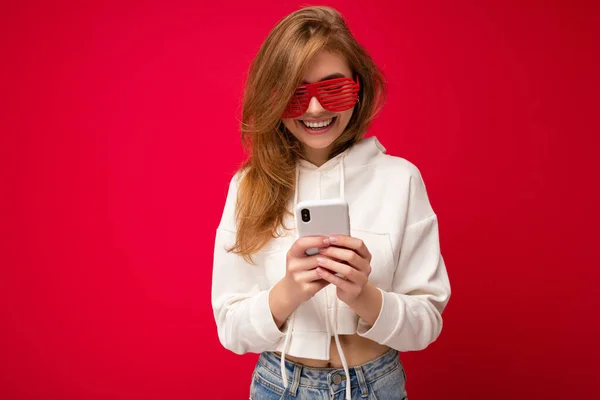 Φωτογραφία από ελκυστική θετική εμφάνιση νεαρή γυναίκα φορώντας casual κομψό ντύσιμο poising απομονωμένη στο παρασκήνιο με κενό χώρο που κατέχουν στο χέρι και τη χρήση sms κινητό τηλέφωνο κοιτάζοντας — Φωτογραφία Αρχείου