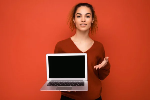 Beautiful charming fascinating young curly dark blond woman wearing red sweater standing isolated over red wall background holding computer laptop with empty copy space mock up pointing to the camera — 图库照片