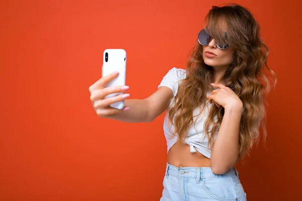 Close-up foto van verbazingwekkende mooie jonge blonde vrouw met mobiele telefoon het nemen van selfie foto met behulp van smartphone camera dragen zonnebril dagelijks stijlvolle outfit geïsoleerd over kleurrijke muur achtergrond — Stockfoto