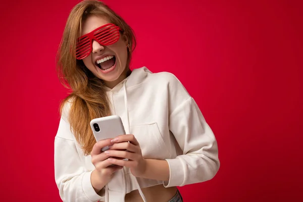 Overjoyed young blonde woman wearing stylish white hoodie and funny colorful glasses standing isolated over colourful background holding and using mobile phone communicating online on the internet — Stock Photo, Image
