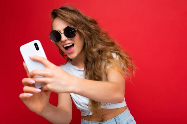 Ελκυστική γοητευτική νεαρή χαμογελαστή ευτυχισμένη γυναίκα κρατώντας και χρησιμοποιώντας το κινητό τηλέφωνο λήψη selfie φορώντας κομψά ρούχα απομονώνονται σε φόντο τοίχο — Φωτογραφία Αρχείου
