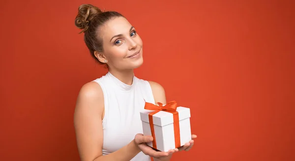 Elég boldog fiatal szőke nő személy elszigetelt felett piros háttér fal visel fehér felső gazdaság ajándék doboz és nézi a kamera — Stock Fotó
