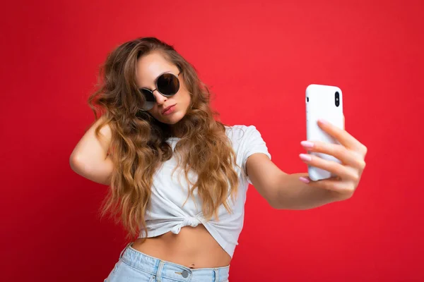 Primer plano de la hermosa mujer hermosa sexy sosteniendo el teléfono móvil tomando foto selfie usando la cámara del teléfono inteligente usando gafas de sol redondas conjunto elegante todos los días aislado sobre fondo de pared colorido —  Fotos de Stock