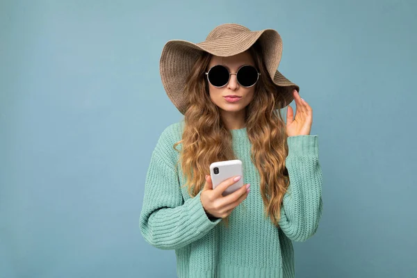 Piękna młoda blondynka w niebieskim swetrze kapelusz i okulary przeciwsłoneczne stojące na niebieskim tle surfing w Internecie przez telefon patrząc na gadjet — Zdjęcie stockowe