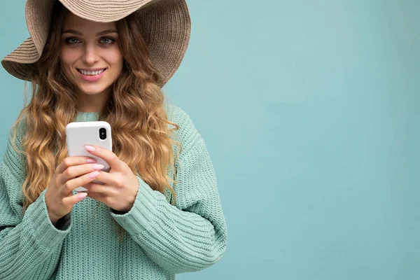 Closeup portrait photo of beautiful young blonde woman wearing blue sweater and hat standing isolated over blue background surfing on the internet via phone looking at camera — Fotografia de Stock