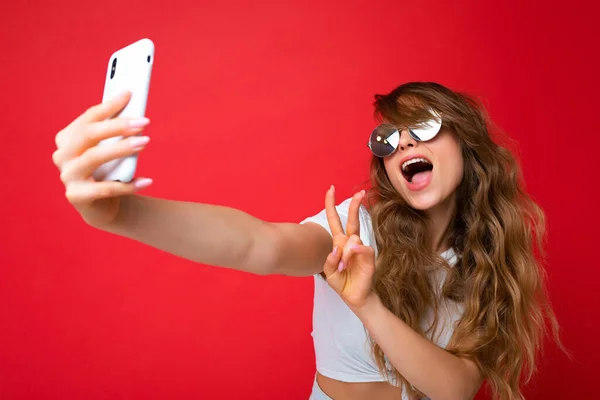 Foto close-up de bela jovem segurando telefone celular tirando foto selfie usando câmera de smartphone usando óculos de sol roupa elegante todos os dias isolado sobre fundo de parede colorido olhando para — Fotografia de Stock