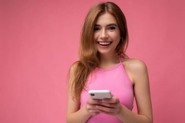 Pembe bluz giyen güzel sarışın bir kadının fotoğrafı. Pembe arka planda izole edilmiş. Elinde boş bir alan var ve kameraya bakan cep telefonu mesajları kullanıyor.