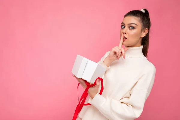 Attraktiv positiv överraskad ung brunett kvinna isolerad över rosa bakgrund vägg bär vit tröja innehav presentförpackning och unboxing närvarande tittar på kameran och visar shh gest — Stockfoto