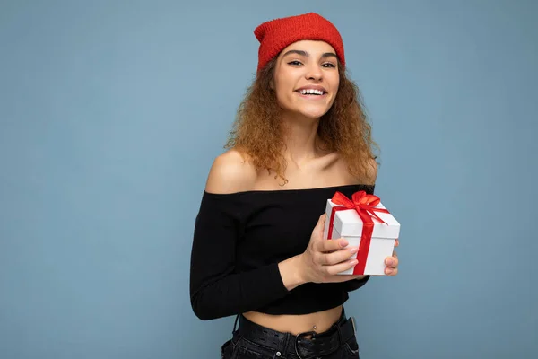 Tiro de atractiva mujer joven sonriente positiva aislada sobre la pared de fondo colorido que lleva traje de moda todos los días sosteniendo la caja de regalo y mirando a la cámara — Foto de Stock