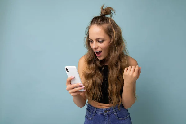 Charming positive young blonde woman wearing black top poising isolated on blue background with empty space holding in hand and using mobile phone communicating online looking at gadjet — Fotografia de Stock