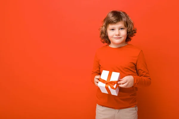 Tiro de niño rizado rubio positivo feliz de pie aislado sobre la pared de fondo rojo con suéter rojo que sostiene la caja de regalo y mirando a la cámara — Foto de Stock