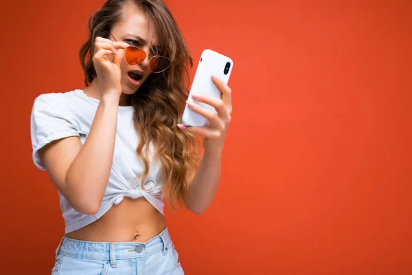 Upset beautiful young woman holding mobile phone wearing sunglasses everyday stylish outfit isolated over wall background looking at device screen and reading news in the internet and dont — 图库照片