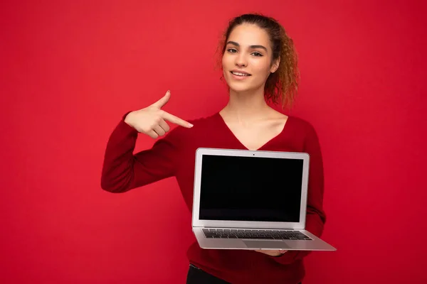 Close-up portrait of beautiful dark blond woman holding laptop computer looking at camera pointing finger at netbook monitor wearing red sweater isolated over red wall background — Φωτογραφία Αρχείου