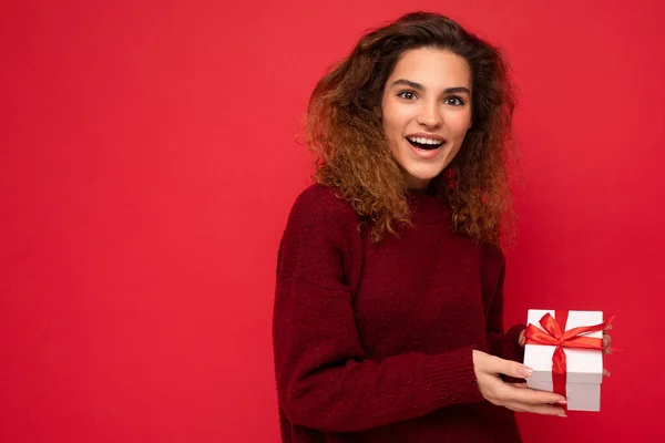 Attractive positive surprised young brunette curly woman isolated over red background wall wearing red sweater holding gift box looking at camera