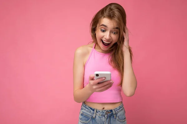 Charmante positieve jonge blonde vrouw dragen roze top vergiftigen geïsoleerd op roze achtergrond met lege ruimte in de hand en met behulp van mobiele telefoon communiceren online kijken naar gadjet — Stockfoto