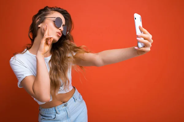 Sexy hermosa mujer rubia joven sosteniendo el teléfono móvil tomando foto selfie usando la cámara del teléfono inteligente usando gafas de sol traje elegante todos los días aislado sobre fondo de pared colorido mirando el dispositivo — Foto de Stock