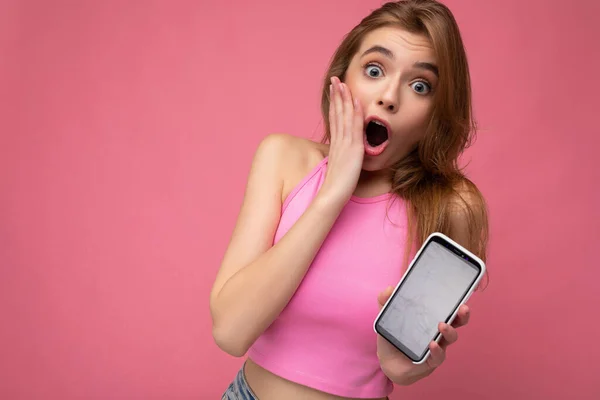 Closeup photo of shocked beautiful young blonde woman wearing pink top poising isolated on pink background with empty space holding in hand and showing mobile phone with empty display for mockup — ストック写真