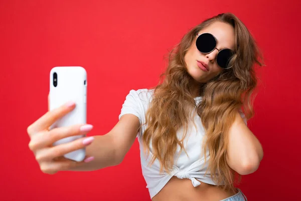 Foto de primer plano de la increíble hermosa mujer rubia joven sosteniendo el teléfono móvil tomando foto selfie utilizando la cámara del teléfono inteligente con gafas de sol traje elegante todos los días aislado sobre fondo de pared colorido —  Fotos de Stock