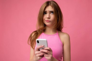 Pembe bluz giyen güzel, düşünceli genç bir kadının yakın plan fotoğrafı. Pembe arka planda izole edilmiş, el ele tutuşan ve internette cep telefonu sörfü yapan bir kadın.