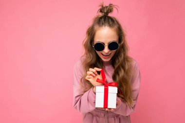 Renkli arka plan duvarından izole edilmiş, şık kıyafetler giymiş, elinde hediye kutusu olan ve içinde kırmızı kurdele olan hediye kutusuna bakan sarışın bir kadının oldukça olumlu bir fotoğrafı.