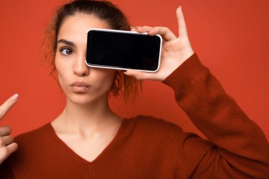 Kırmızı arka plan üzerinde koyu kırmızı süveter giyen neşeli genç bir kadının yakın plan fotoğrafı. Akıllı telefonu tutuyor ve cep telefonu ekranını fotokopi makinesine bakmak için fotokopi alanı ile gösteriyor.