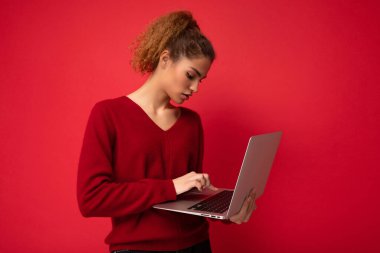 Yakından çekilmiş bir portre, elinde dizüstü bilgisayarla netbook monitörüne bakıp kırmızı duvar arkaplanından izole edilmiş kırmızı kazak giyen güzel sarışın bir kadın.
