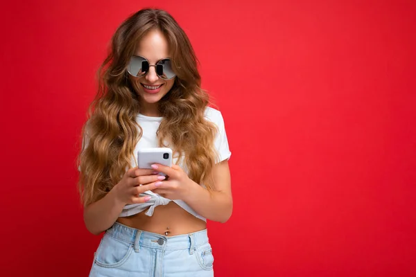 Beautiful young woman wearing casual clothes standing isolated over background surfing on the internet via phone looking at mobile screen — Stock Photo, Image