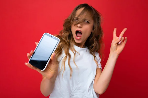 Foto von schönen erstaunt erstaunt junge blonde Frau gut aussehend trägt weißes T-Shirt isoliert auf rotem Hintergrund mit Kopierraum hält Telefon zeigt Smartphone in der Hand mit leeren — Stockfoto