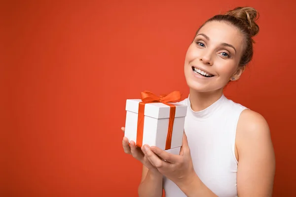 아름답고 행복 한 젊은 금발 여성 이 선물 상자를 들고 카메라를 보며 화려 한 평상복을 입고 다채 로운 배경 벽에 고립되어 있다 — 스톡 사진
